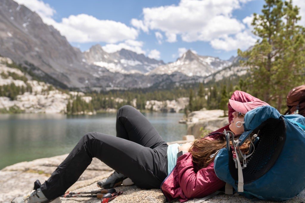 woman sleeping during the day on her backpack by a lake in the mountains
